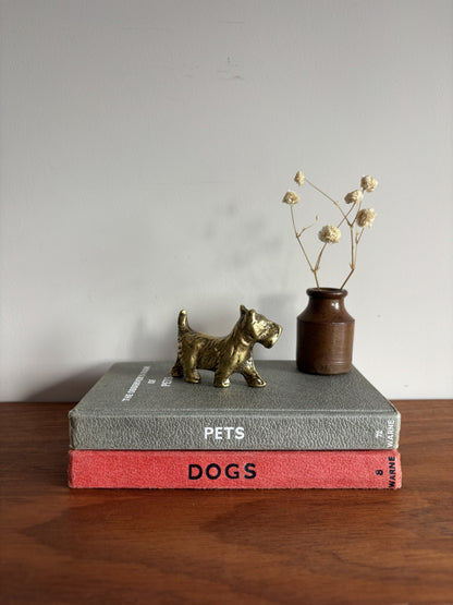 A small brass Scottish Terrier figurine stands on top of two stacked books, with a small brown vase holding dried stems beside it, set on a wooden surface against a plain light wall.