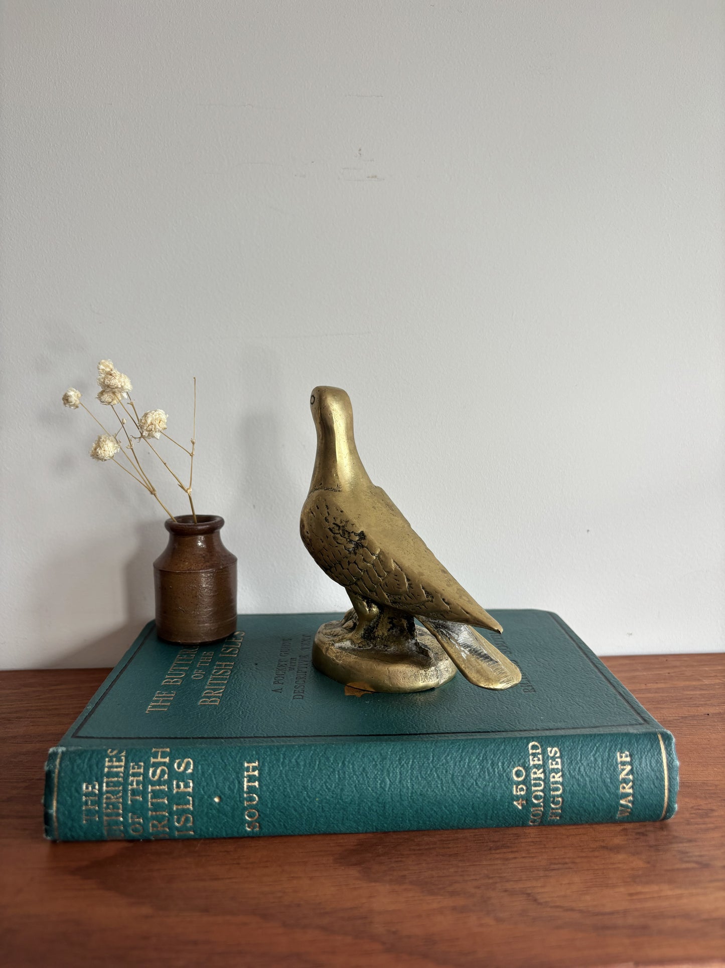A brass dove or pigeon figurine standing on a round base, placed on top of a teal book. A small brown vase with dried stems sits beside it on a wooden surface against a plain light wall.