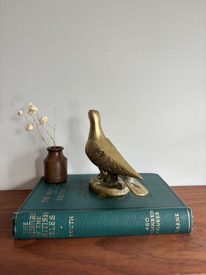 A brass dove or pigeon figurine standing on a round base, placed on top of a teal book. A small brown vase with dried stems sits beside it on a wooden surface against a plain light wall.
