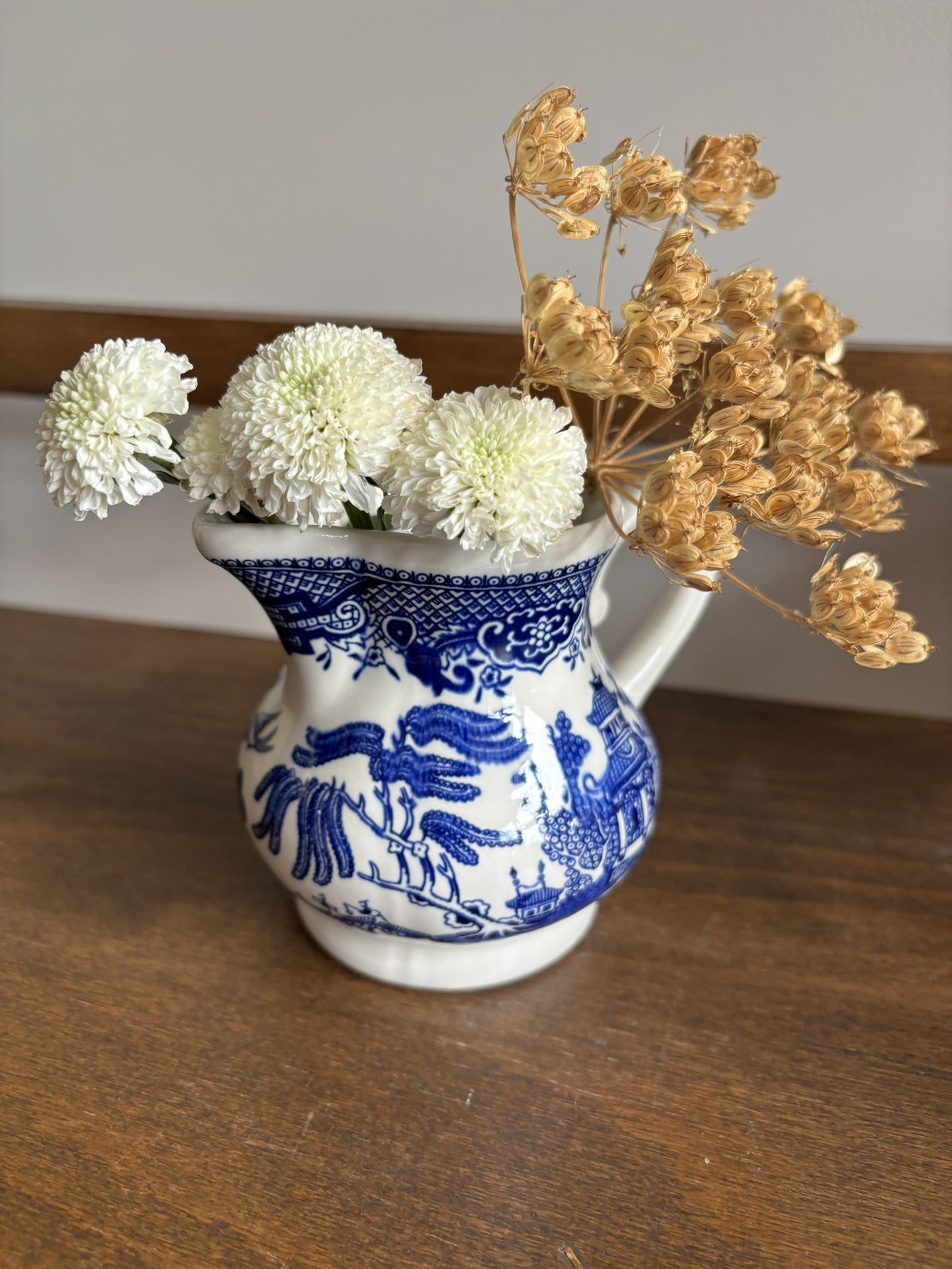 A small white ceramic pitcher with blue floral and bird illustrations, filled with three white pom-pom flowers and dried tan flowers, set on a wooden tabletop against a neutral background.