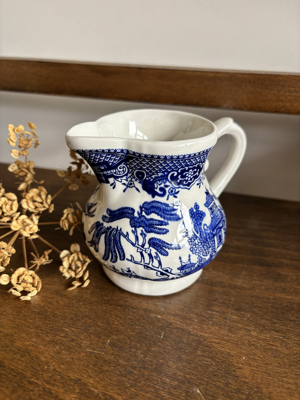 A small white ceramic pitcher with blue floral and landscape illustrations, placed on a wooden surface next to dried flowers.