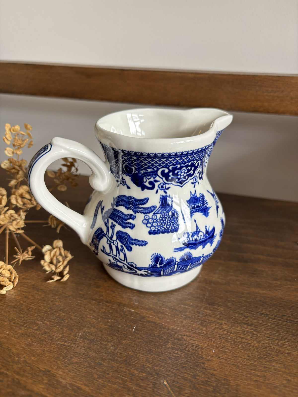 A small white ceramic pitcher with a blue traditional landscape pattern, sitting on a wooden surface next to dried flowers.