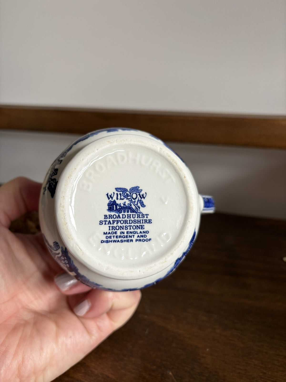 A hand holding the bottom of a white ceramic mug or cup with blue printed branding. The base reads 'WILLOW BROADBURST Staffordshire Ironstone' and shows blue decorative detailing around the rim.