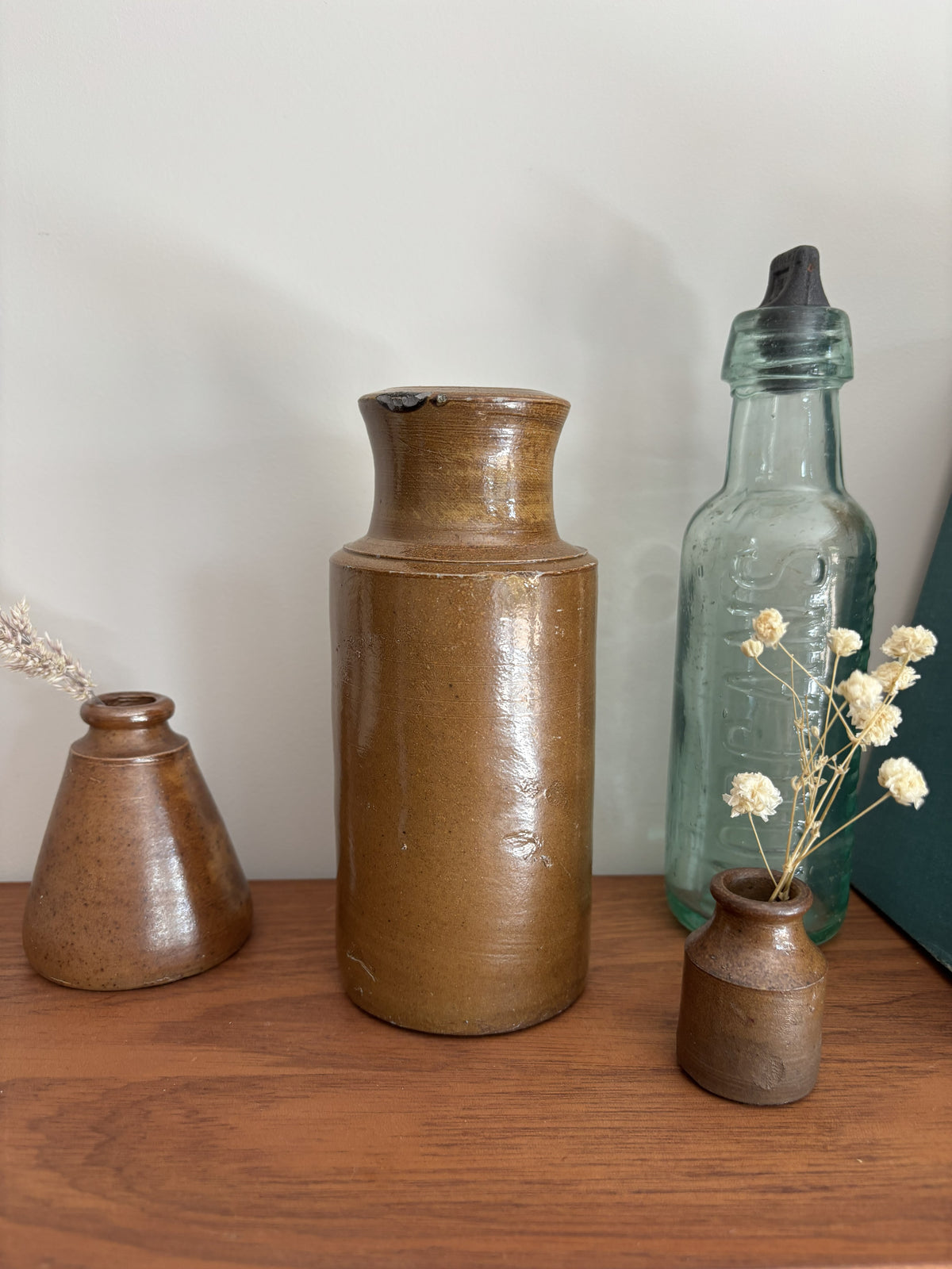 A brown vintage salt glazed ink pot arranged on a wooden surface against a plain light wall. The center vase is tall and cylindrical with a glossy brown glaze, flanked by a smaller brown vase on the left and a translucent pale blue-green bottle with a black stopper on the right.
