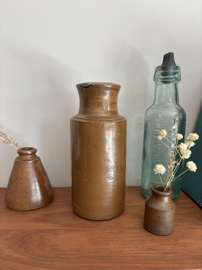 A brown vintage salt glazed ink pot arranged on a wooden surface against a plain light wall. The center vase is tall and cylindrical with a glossy brown glaze, flanked by a smaller brown vase on the left and a translucent pale blue-green bottle with a black stopper on the right.
