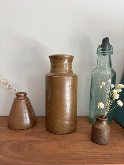 Three rustic ceramic vases in earthy brown tones displayed on a wooden surface against a light gray wall, alongside a vintage glass bottle and dried white flowers.