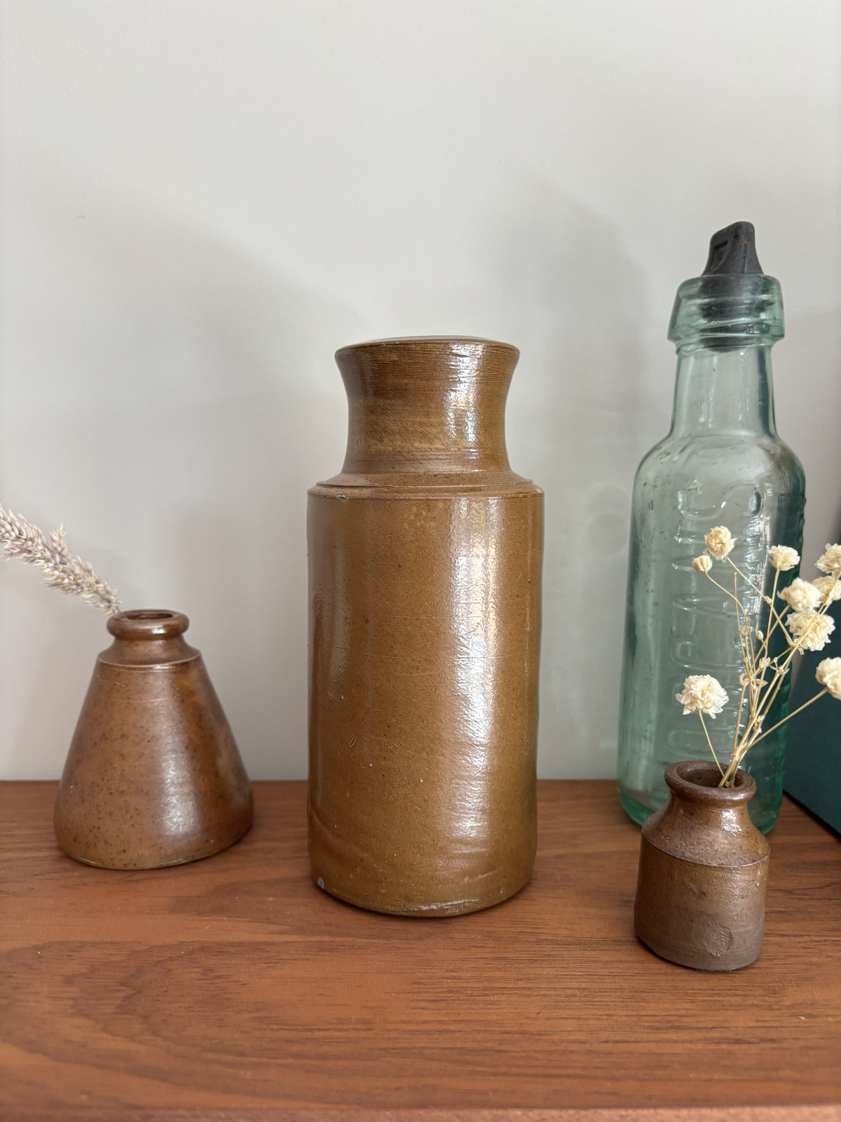 Three rustic ceramic vases in earthy brown and bronze glazes arranged on a wooden shelf, alongside a vintage-style clear glass bottle and dried white flowers, against a plain light gray wall.