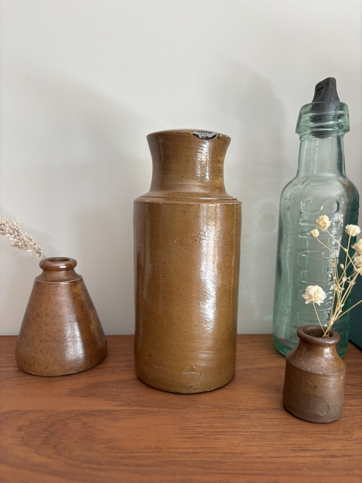 Three rustic ceramic and glass vases displayed on a wooden surface against a plain light wall. The center vase is a tall cylindrical brown ceramic vessel with a glossy finish.