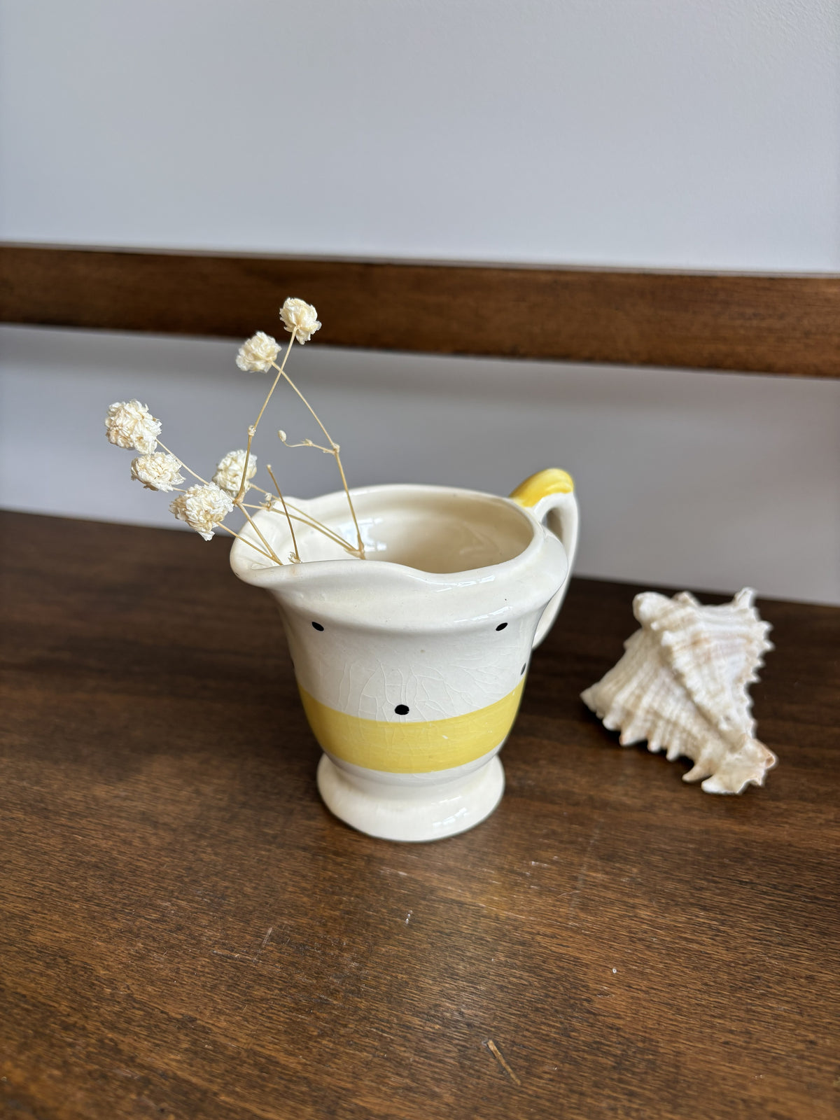 A small ceramic pitcher with a yellow band and black polka dots sits on a wooden table next to a seashell, with dried white flowers placed inside the pitcher.