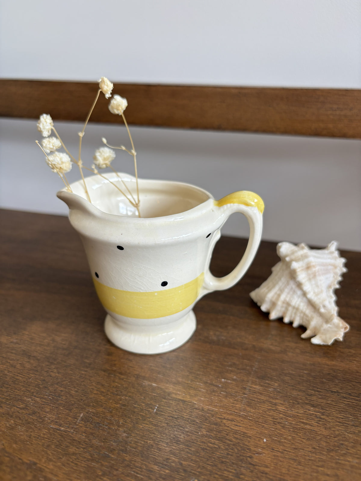 A small cream-colored ceramic pitcher with a yellow band and black polka dots, styled with dried baby's breath stems and a seashell on a wooden surface.
