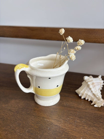 A small cream-colored ceramic pitcher with a yellow band and black polka dots, styled with dried baby's breath stems and a seashell on a wooden surface.
