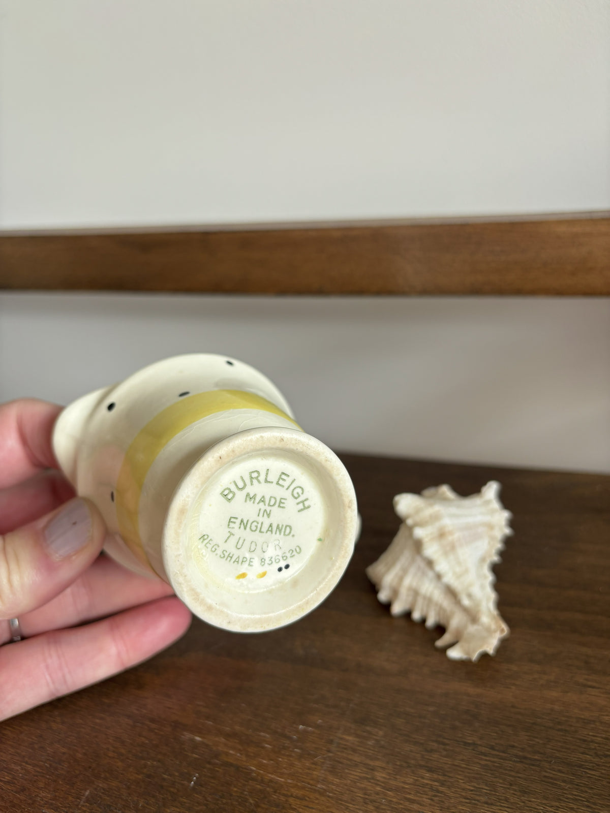 A hand holds a small cream-colored ceramic cup or candle holder showing the printed base, which reads 'BURLEIGH MADE IN ENGLAND'. A seashell rests on a wooden table next to it, with a wooden shelf and light-colored wall in the background.