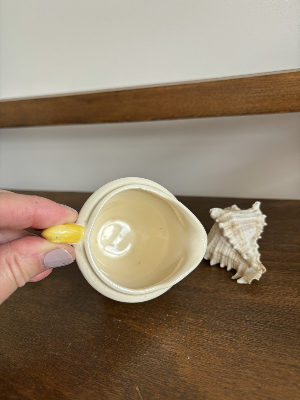 A hand holds a small cream-colored ceramic pitcher with a yellow handle and spout, next to a seashell on a dark wooden surface.