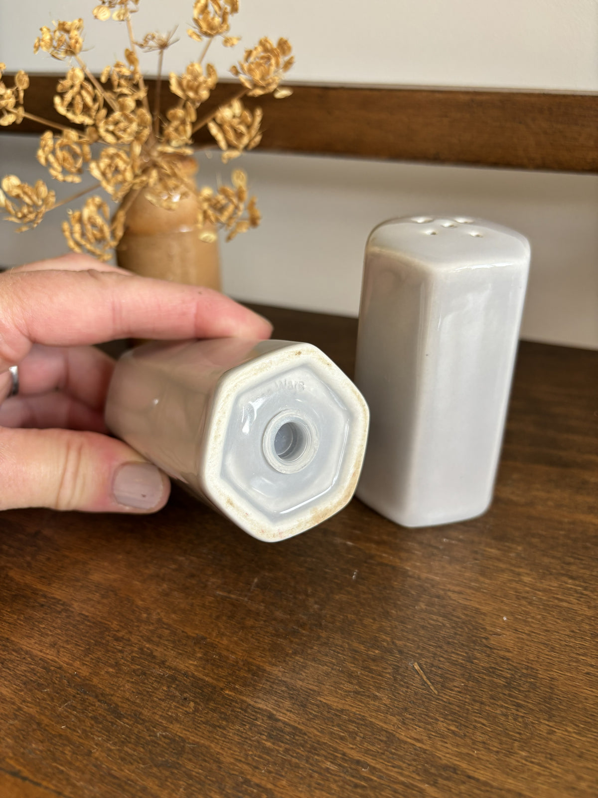 A hand holds a ceramic salt or pepper pot upside down to show the bottom opening, with a matching cylindrical container standing beside it on a wooden table.