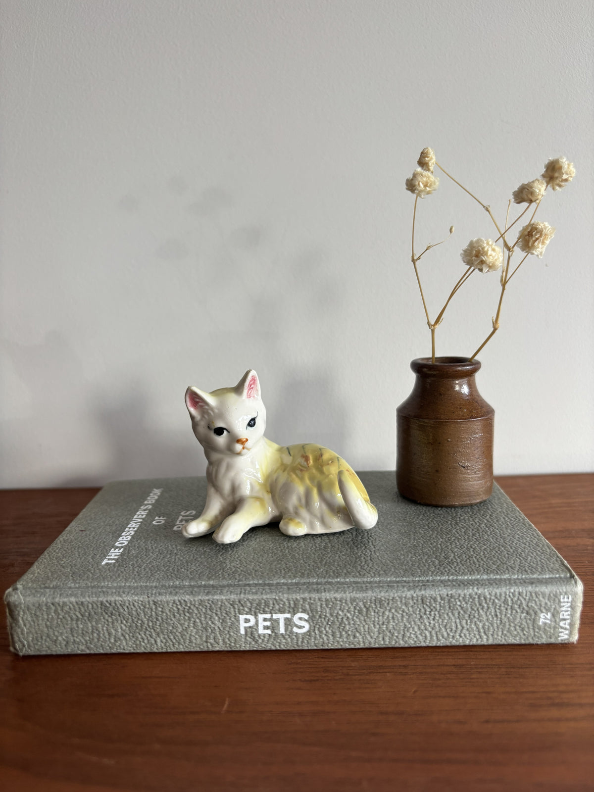 A small white and yellow cat figurine posed on top of a gray book, next to a brown ceramic vase holding dried stems, set on a wooden surface against a plain light wall.