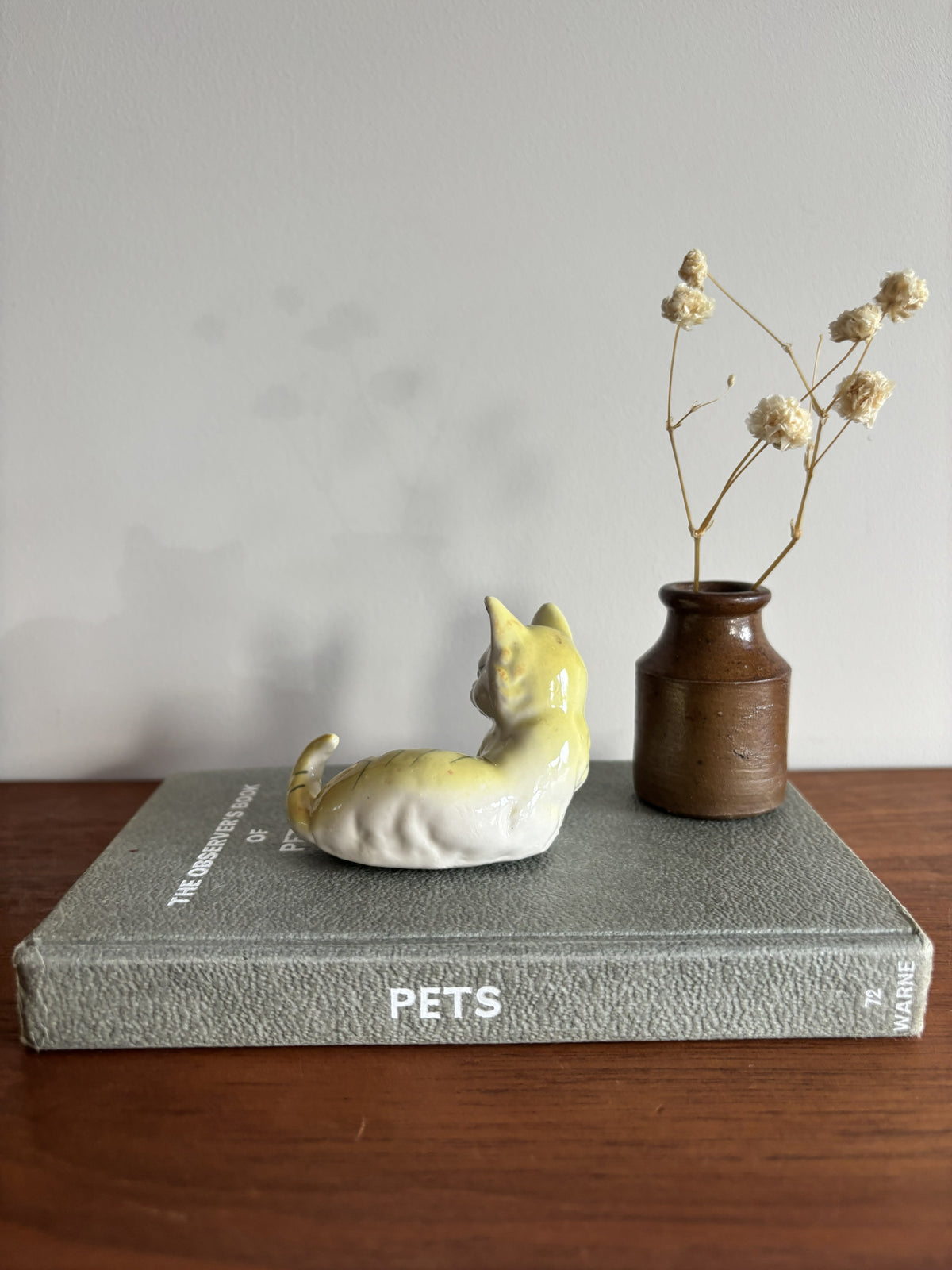A ceramic cat figurine in a yellow-and-white glossy finish, posed sitting on a gray textured book. Next to it is a small brown ceramic vase holding dried cream-colored flowers.