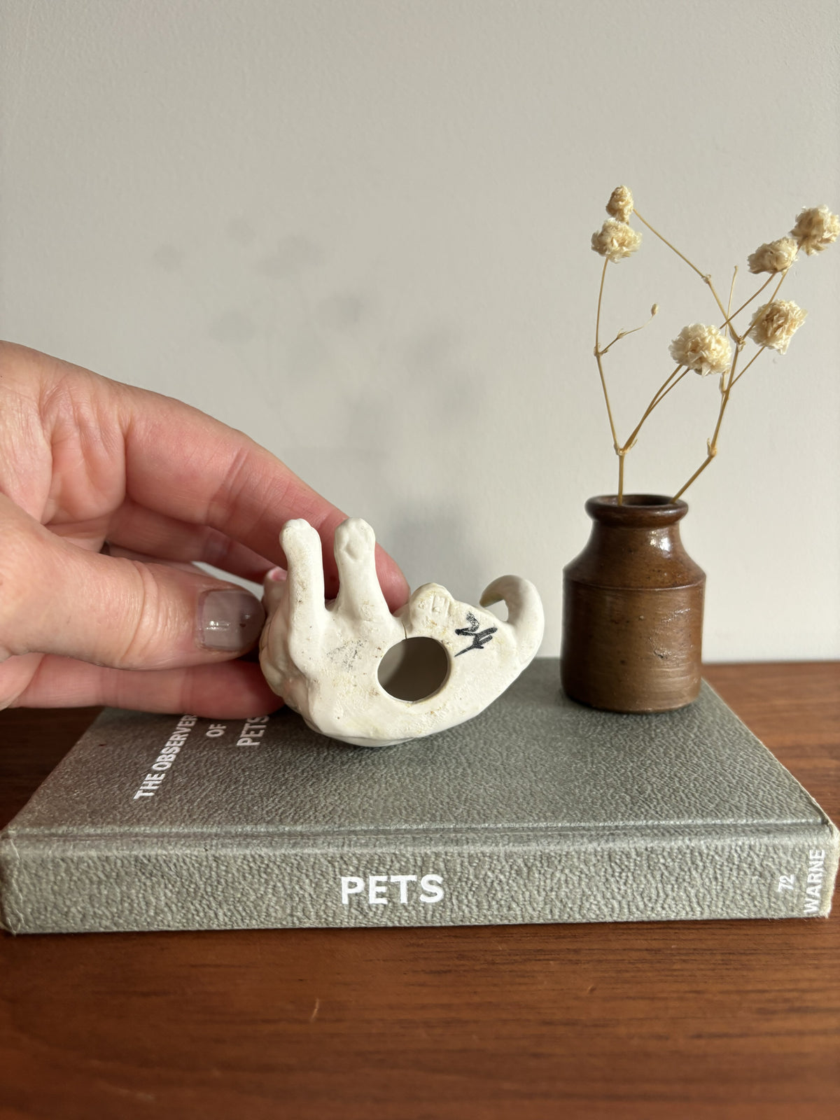 A hand holds a small white ceramic cat-shaped ornament with a circular hole, resting on a gray book. A small brown ceramic vase with dried stems stands beside it on a wooden surface against a light wall.