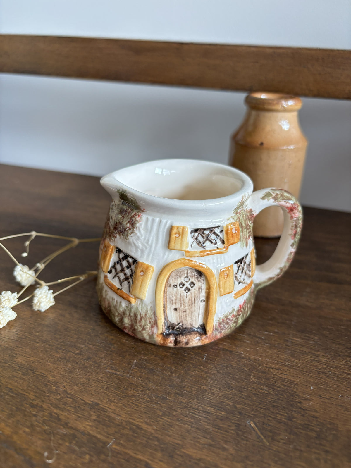 A ceramic cottage-shaped creamer pitcher with a white interior and a hand-painted exterior featuring a brown door, yellow windows, and a rustic floral pattern, set on a wooden table.