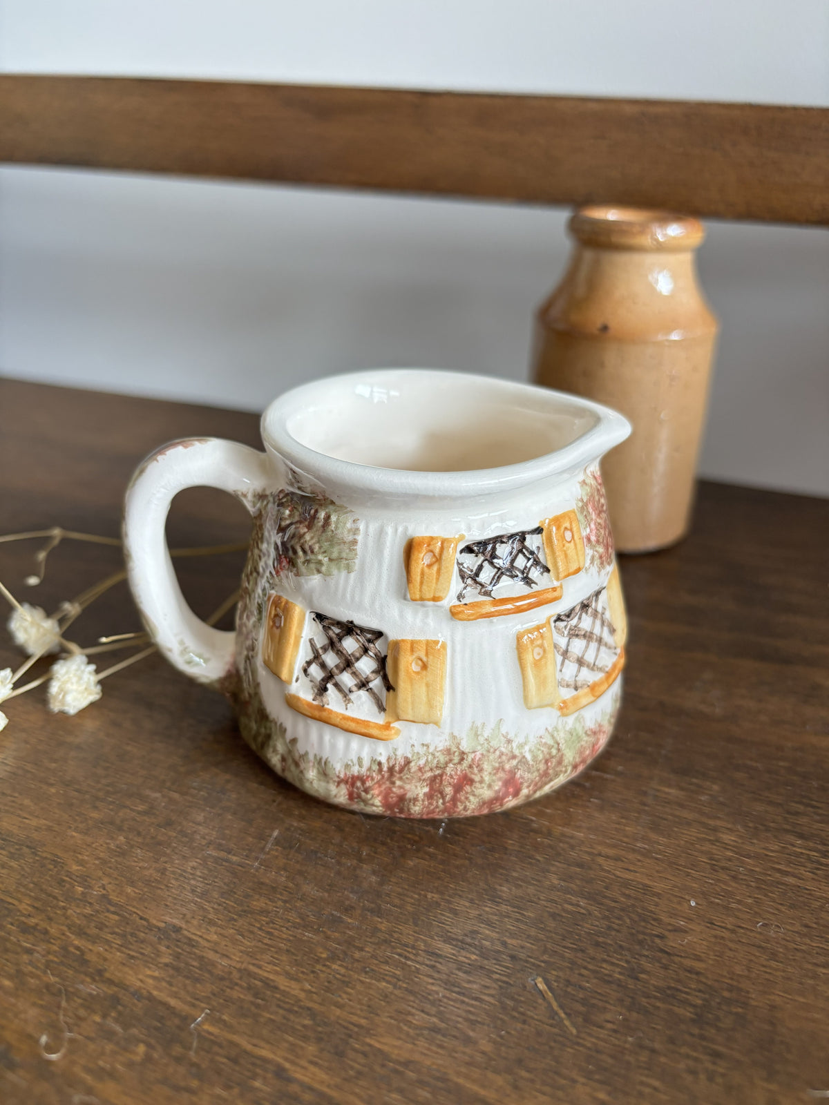 A small ceramic pitcher with a handle, decorated with a rustic pattern of yellow window shutters and lattice windows on a white base, sitting on a wooden table with a blurred brown bottle in the background.