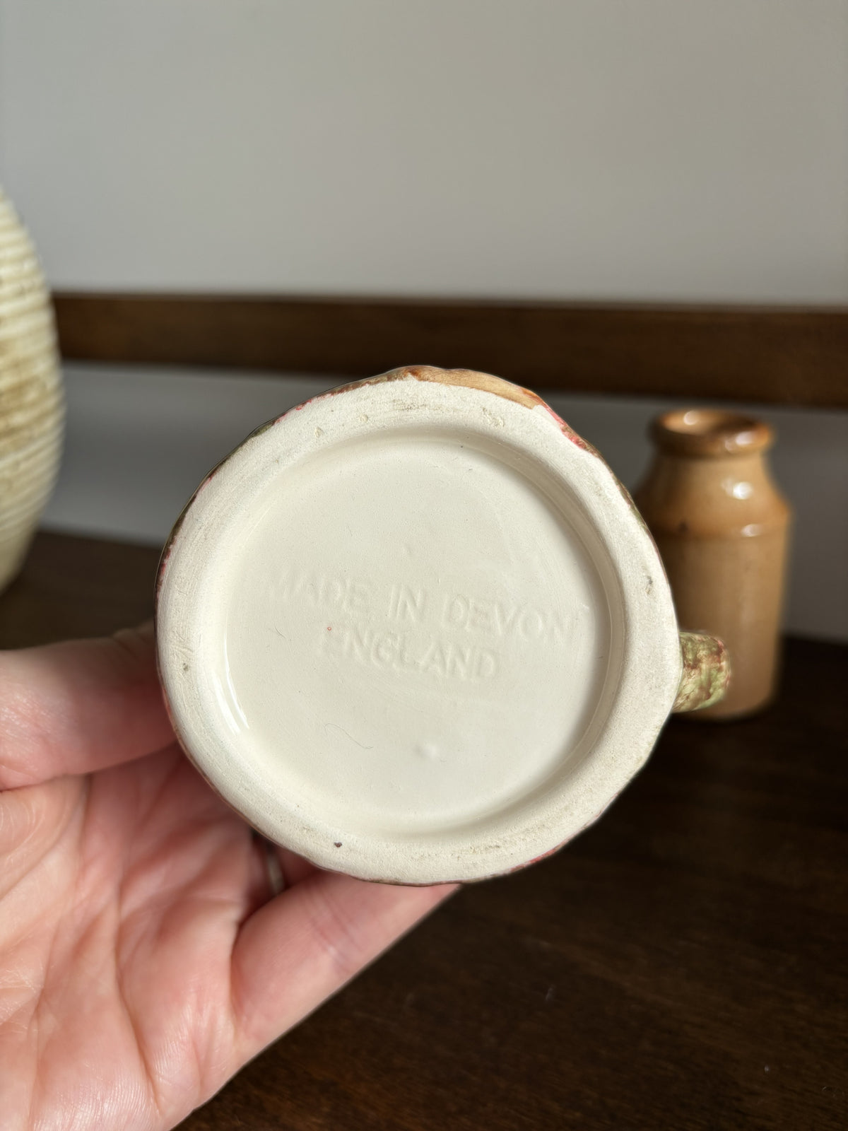 A hand holding the bottom of a ceramic mug or bowl, showing the cream-colored glazed base with embossed text reading 'MADE IN DEVON ENGLAND'. The background shows a dark wooden table and a small brown bottle.