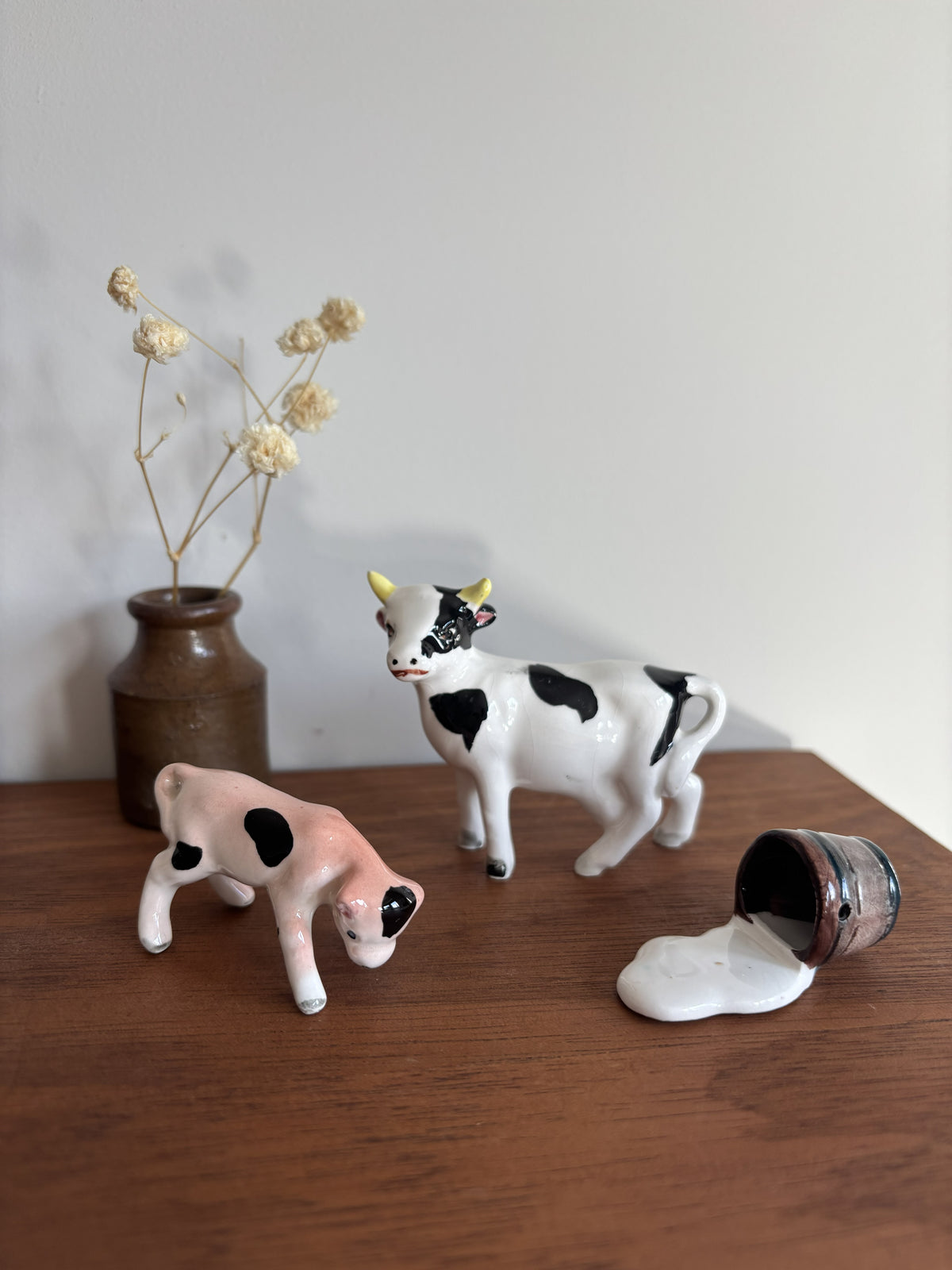 Two ceramic cow figurines on a wooden tabletop against a plain wall, with a small brown vase of dried flowers behind them and a tipped-over bucket with spilled white liquid in the foreground.
