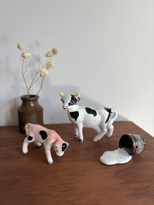 Two ceramic cow figurines on a wooden tabletop against a plain wall, with a small brown vase of dried flowers behind them and a tipped-over bucket with spilled white liquid in the foreground.
