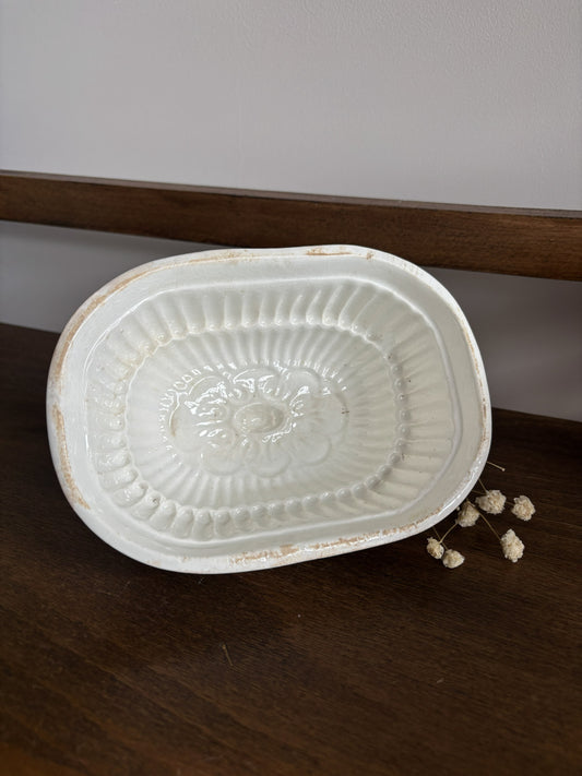 A white ceramic oval jelly mould with a distressed finish and embossed floral detailing in the center, resting on a dark wooden surface.