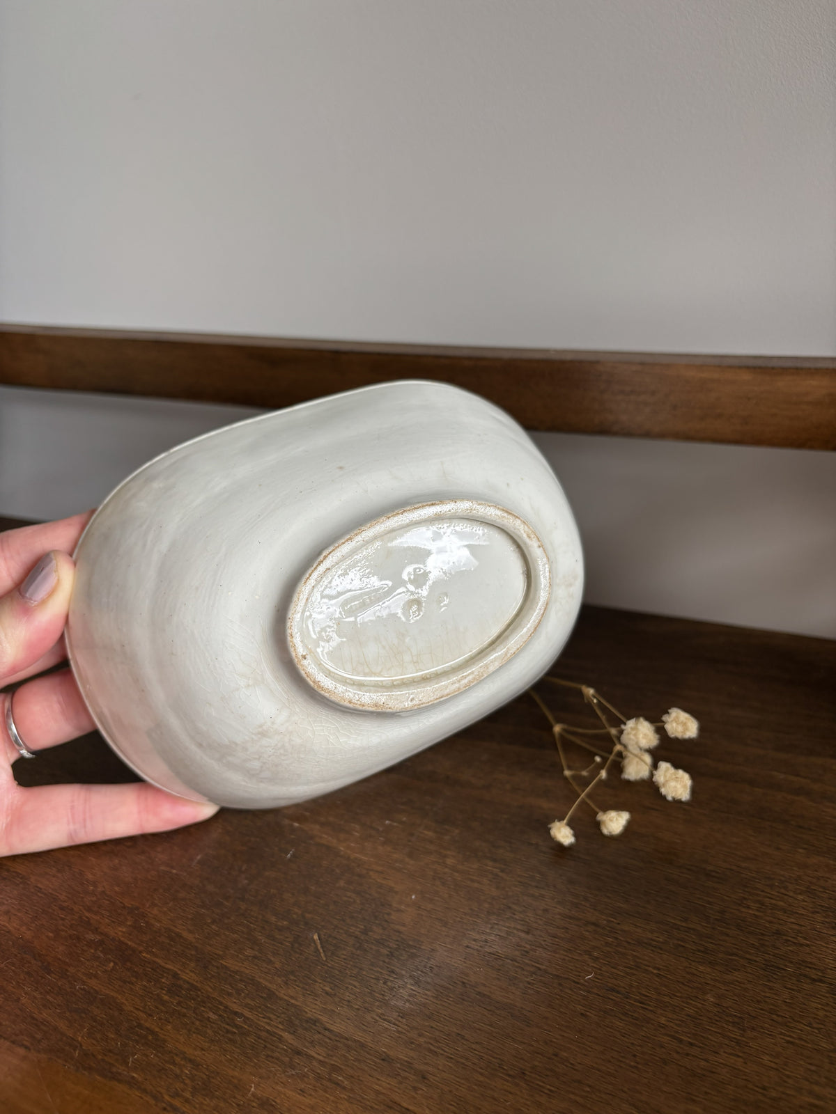 A hand holds a white ceramic jelly mould upside down on a dark wooden table, showing the glazed bottom with a circular foot ring. A small sprig of dried baby's breath lies nearby against a neutral wall background.