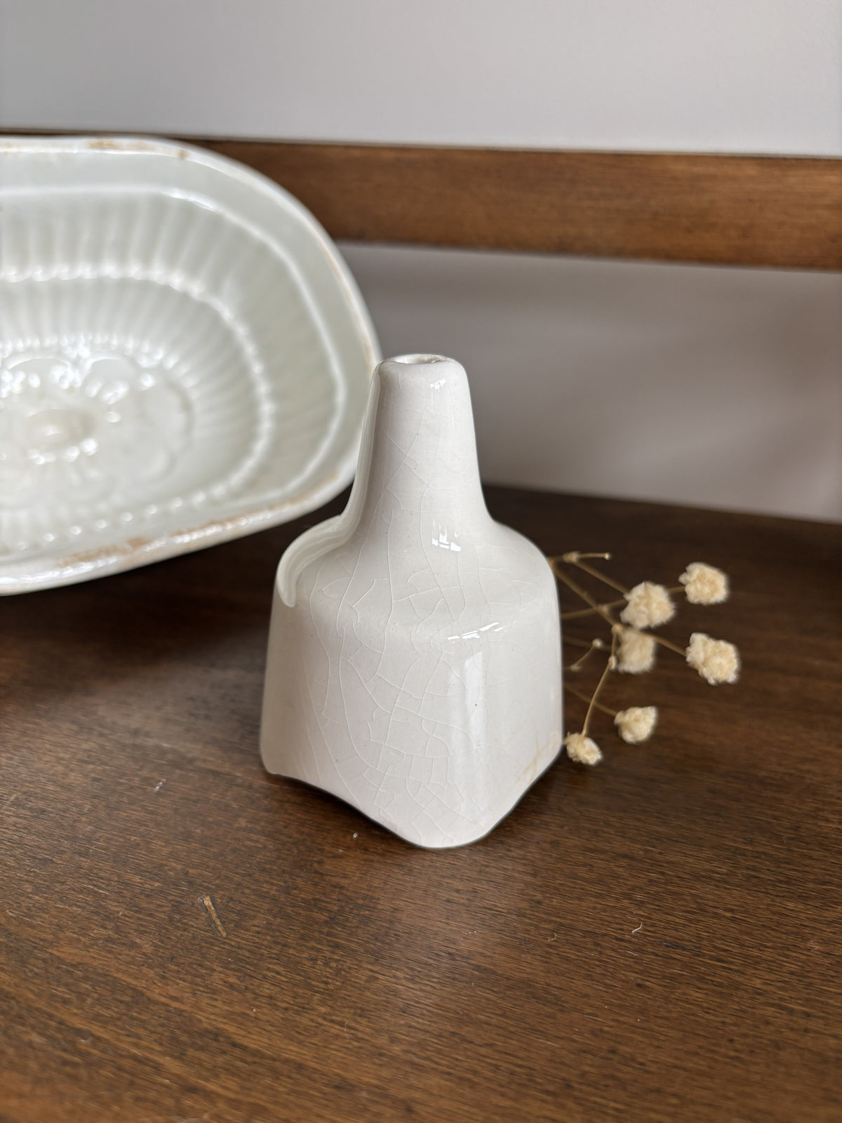 A small white ceramic pie funnel standing on a dark wooden surface next to dried flowers and a matching white ceramic dish in the background.