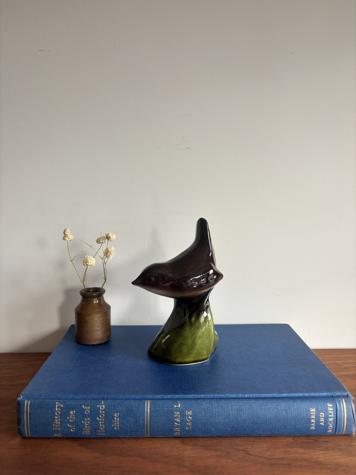 A ceramic bird sculpture with a glossy dark brown and green glaze sits on top of a blue hardcover book on a wooden surface, next to a small brown vase with dried flowers against a plain light gray wall.