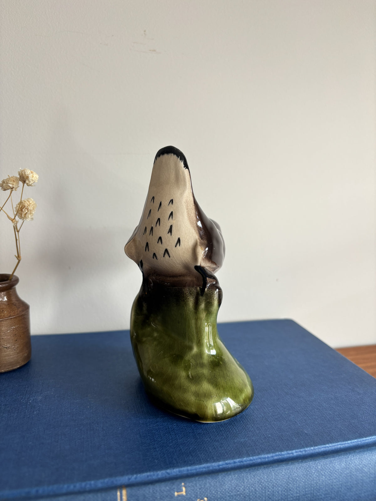 A small glossy ceramic figurine of a bird perched on a green branch, displayed on top of a blue book against a plain light wall, with a small vase of dried stems at the side.