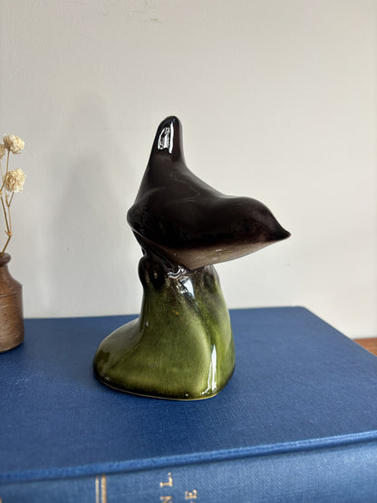 A glossy ceramic bird figurine in a dark brown to olive green glaze, displayed on top of a blue hardcover book against a plain light wall.