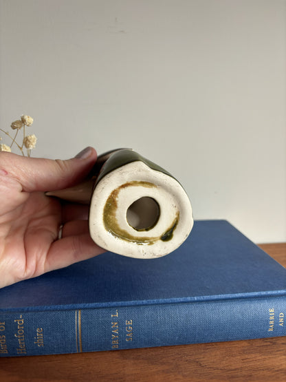 A hand holds a ceramic bird figurine which features a dark green glazed body and a cream-colored interior with a gold-toned ring. The vase is positioned above a blue hardcover book on a wooden surface against a plain light gray wall.