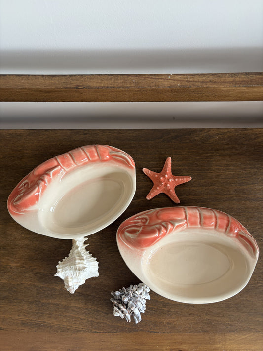 Two oval ceramic dishes with shrimp-shaped rims in a coral pink and cream glaze, styled on a wooden surface with a starfish and seashells.