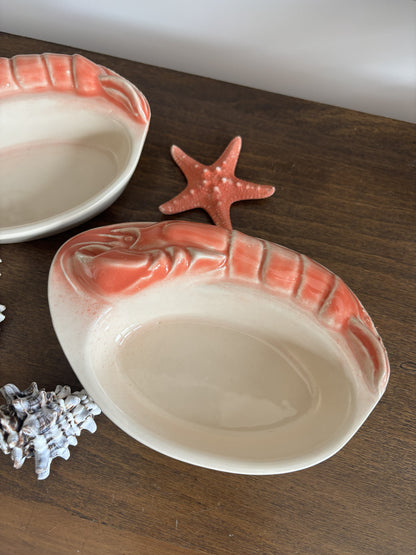 Two ceramic shrimp-shaped serving platters in a warm cream glaze with raised coral-pink shrimp claws, displayed on a dark wood surface with a starfish and seashell props.