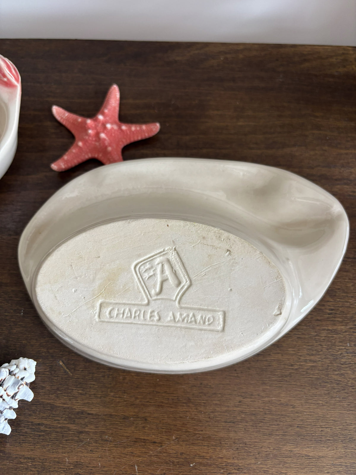 An oval ceramic dish with a raised embossed Charles Amand logo, displayed on a dark wooden surface next to a pink starfish prop.