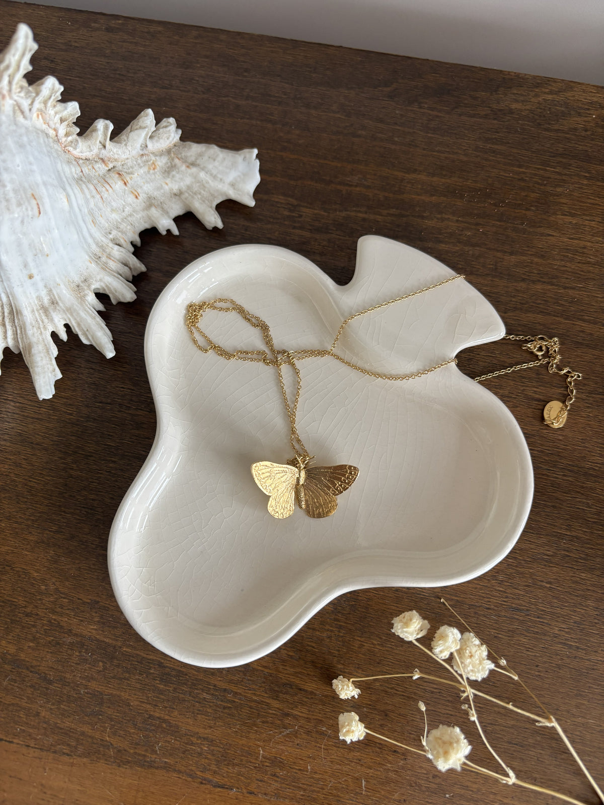 A clover shaped trinket dish holding  butterfly necklace. The scene is styled on a dark wood surface with a white seashell and dried flowers.