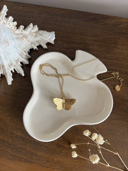 A clover shaped trinket dish holding  butterfly necklace. The scene is styled on a dark wood surface with a white seashell and dried flowers.