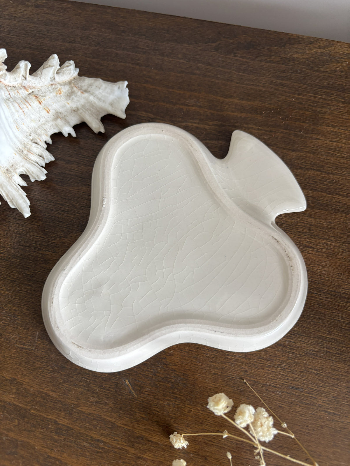 The base of a clover shaped trinket dish holding  a butterfly necklace. The scene is styled on a dark wood surface with a white seashell and dried flowers.