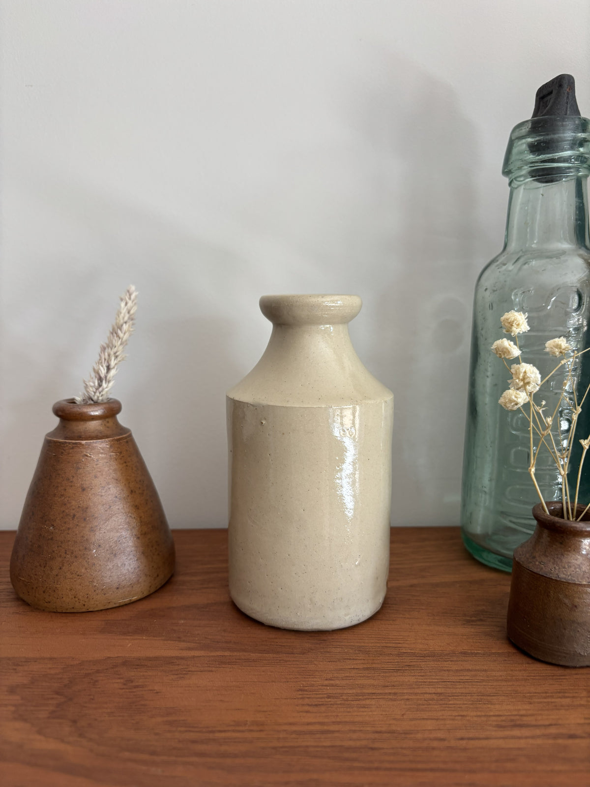 A beige stoneware ink bottle with a glossy finish sits on a wooden shelf, flanked by a brown speckled vase with dried grass on the left and a tall clear glass bottle on the right.