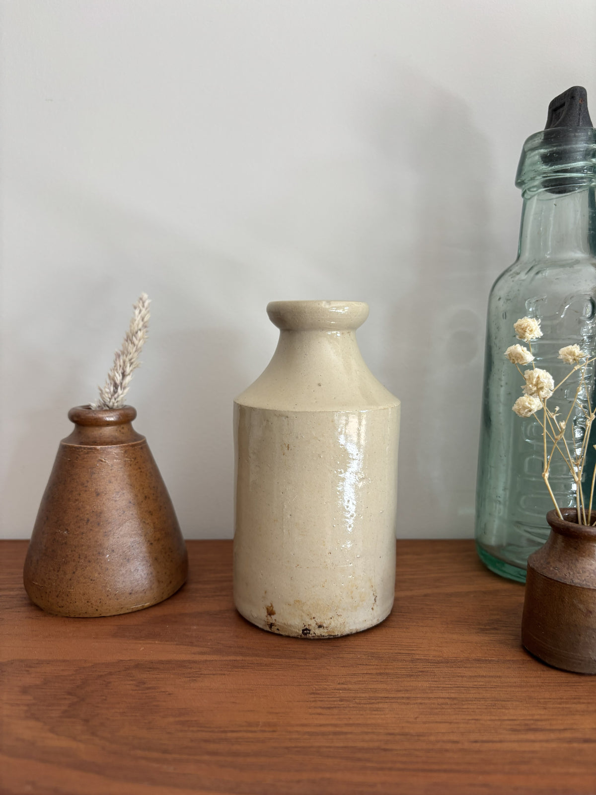 A beige stoneware ink bottle with a glossy finish sits on a wooden shelf, flanked by a brown speckled vase with dried grass on the left and a tall clear glass bottle on the right.