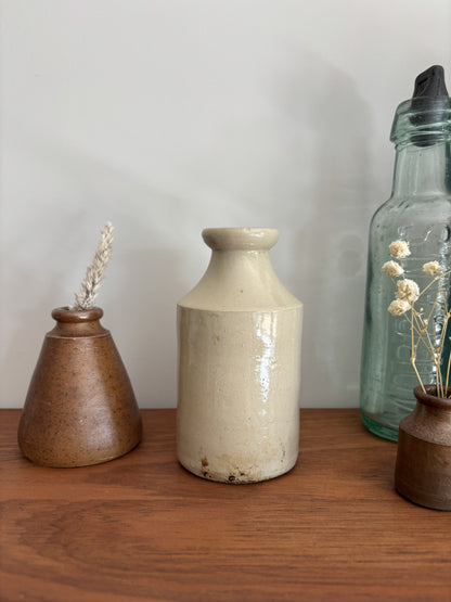 A beige stoneware ink bottle with a glossy finish sits on a wooden shelf, flanked by a brown speckled vase with dried grass on the left and a tall clear glass bottle on the right.