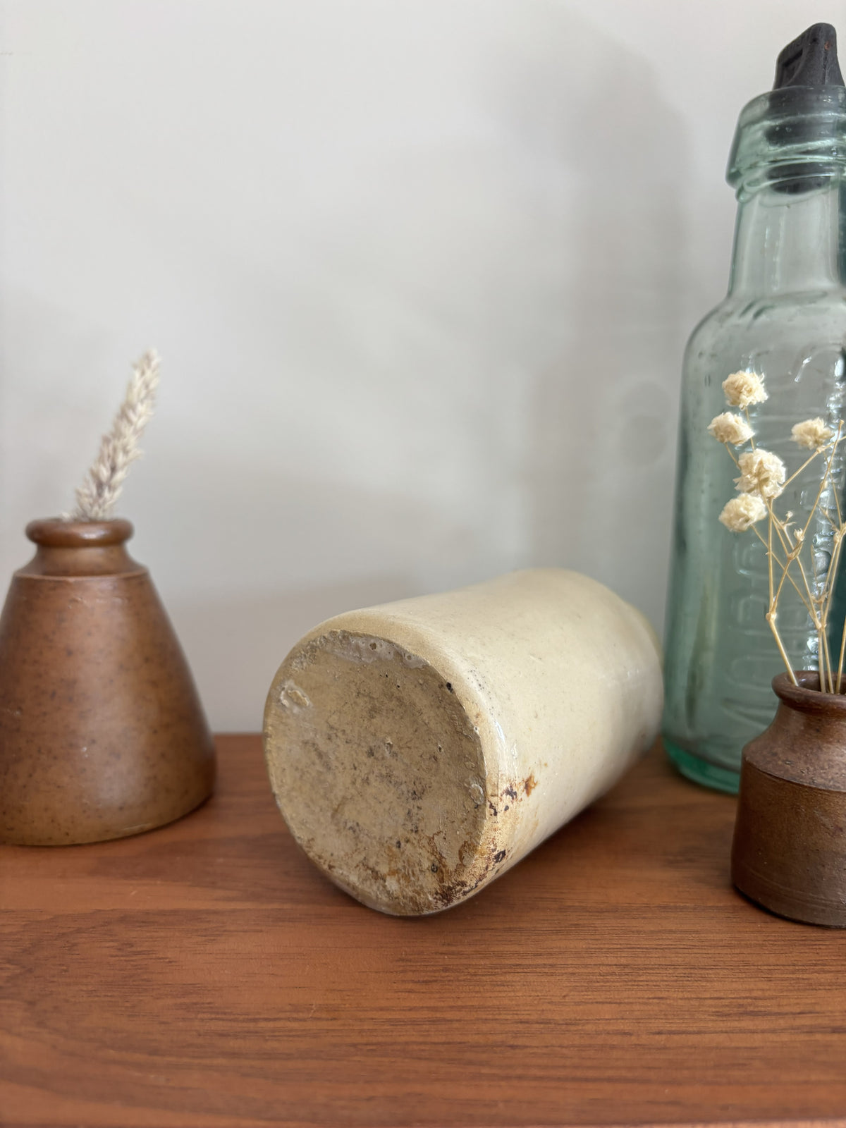 The base of a beige stoneware ink bottle with a glossy finish sits on a wooden shelf, flanked by a brown speckled vase with dried grass on the left and a tall clear glass bottle on the right.