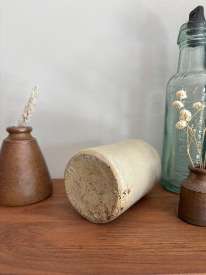 The base of a beige stoneware ink bottle with a glossy finish sits on a wooden shelf, flanked by a brown speckled vase with dried grass on the left and a tall clear glass bottle on the right.