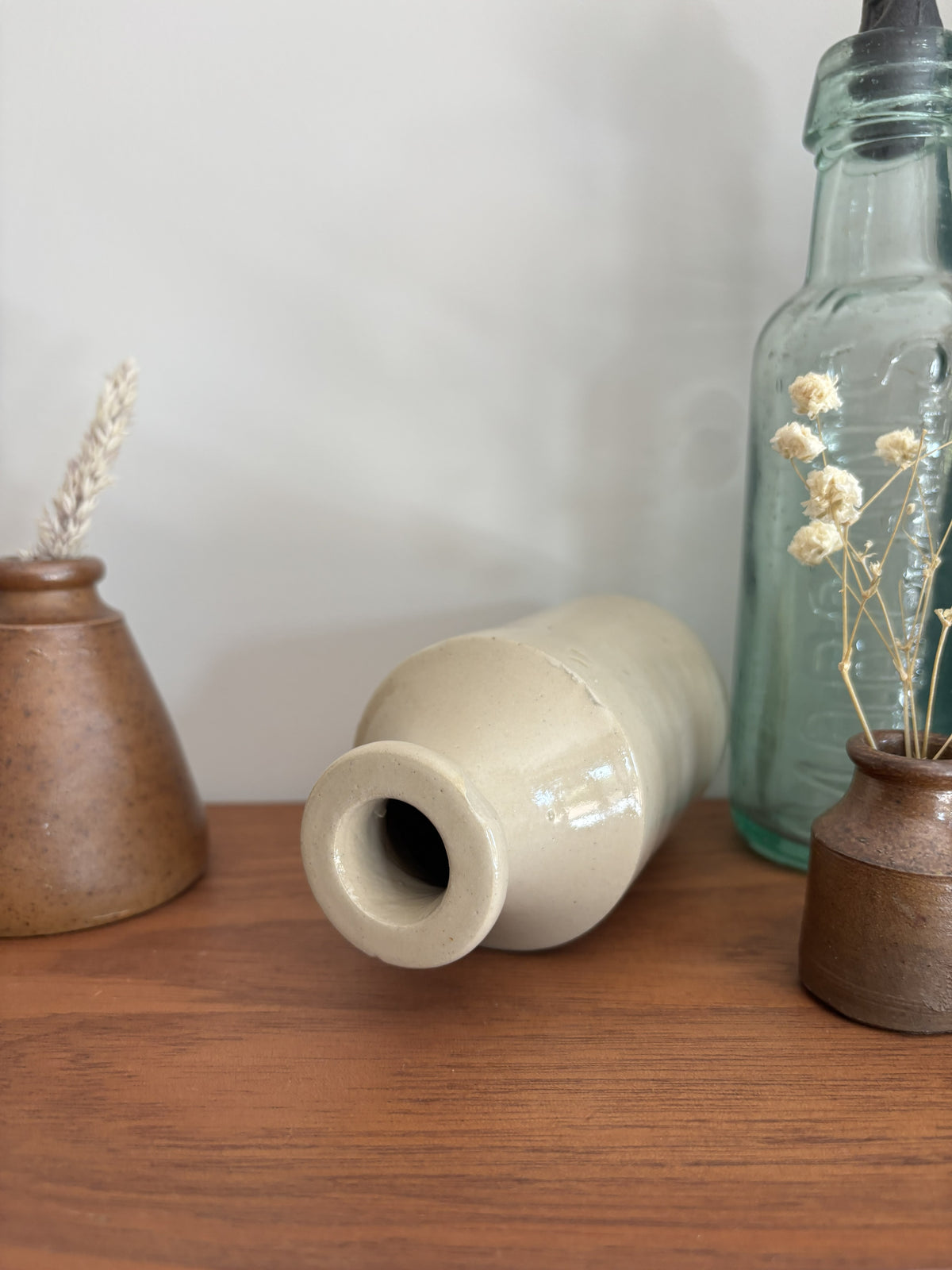 A beige stoneware ink bottle with a glossy finish lying down on a wooden shelf, flanked by a brown speckled vase with dried grass on the left and a tall clear glass bottle on the right.