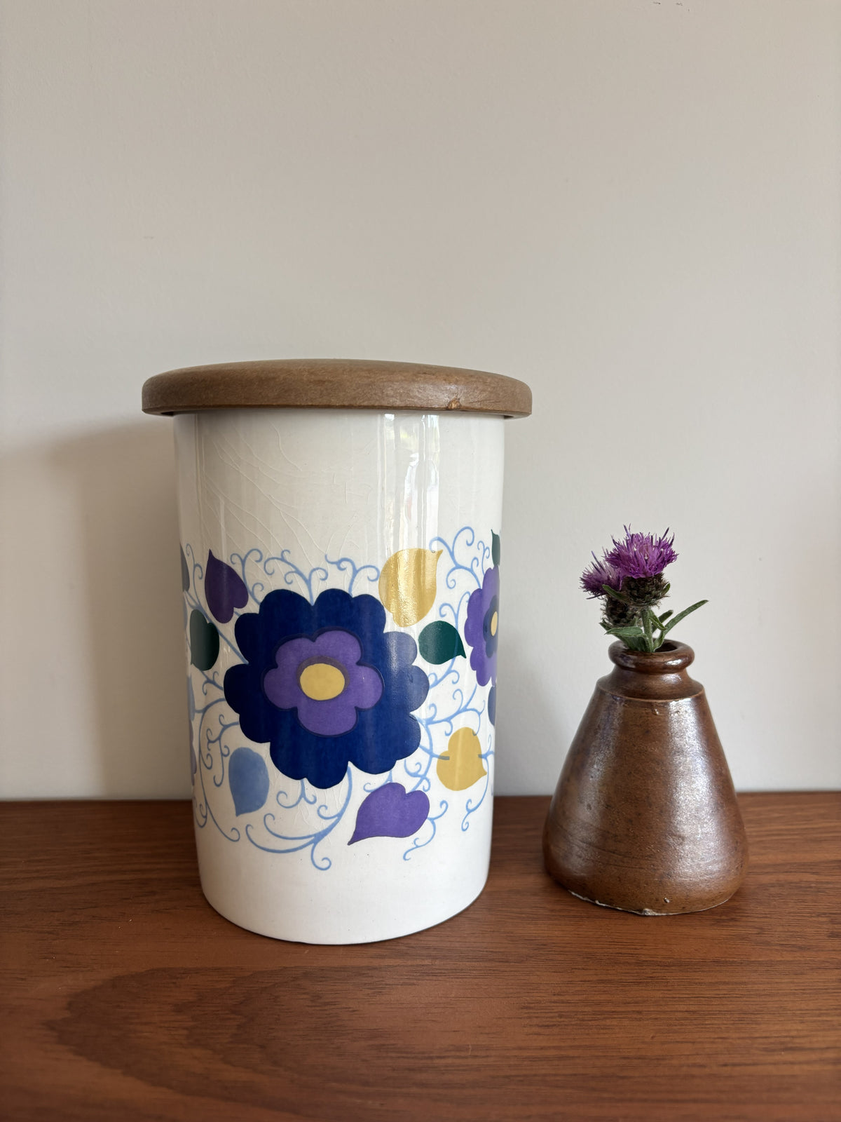 A white Crown Devon ceramic storage jar with a wooden lid, decorated with a blue, purple, and yellow floral pattern.
