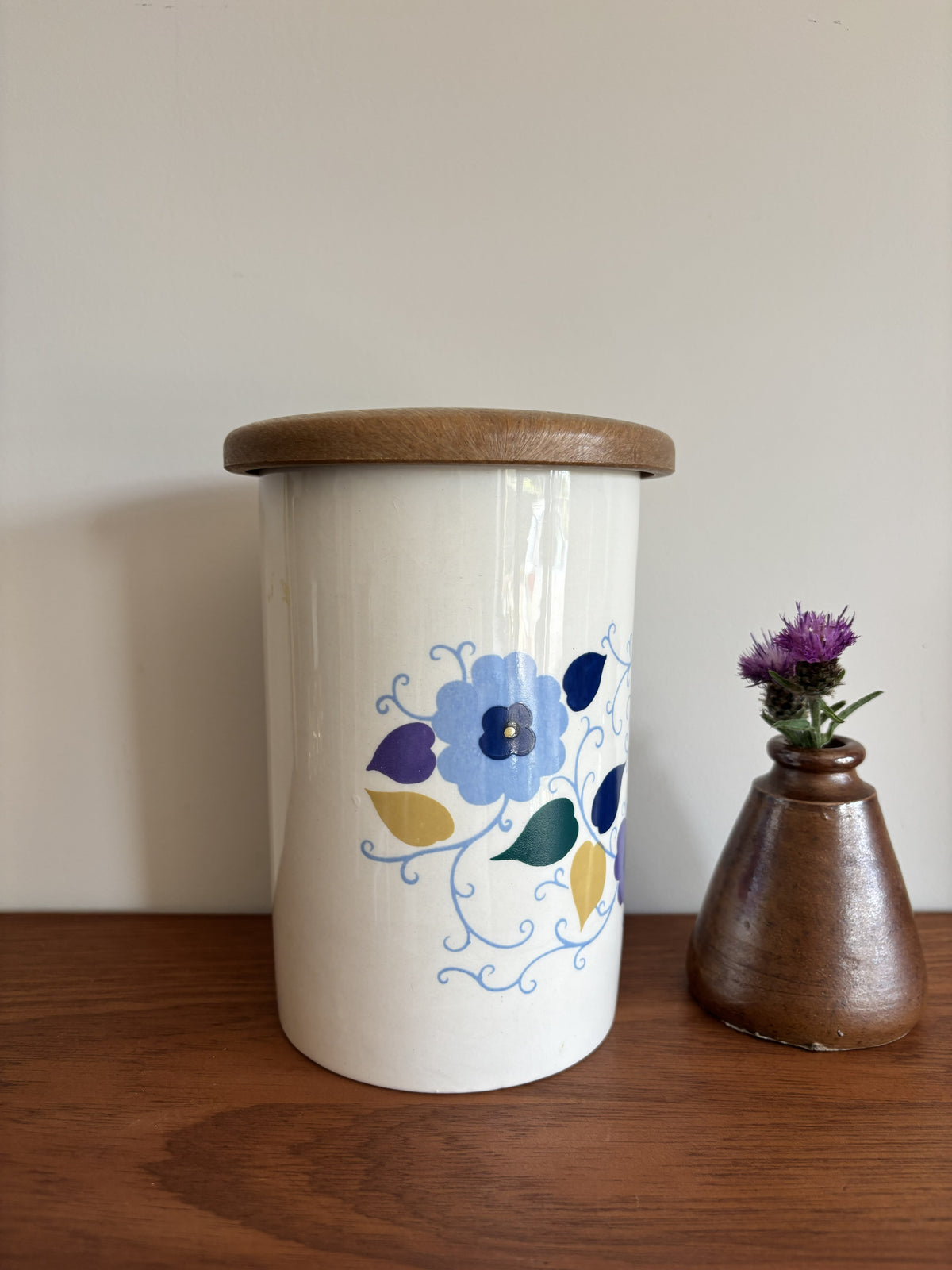 A white Crown Devon ceramic storage jar with a wooden lid, decorated with a blue, purple, and yellow floral pattern.