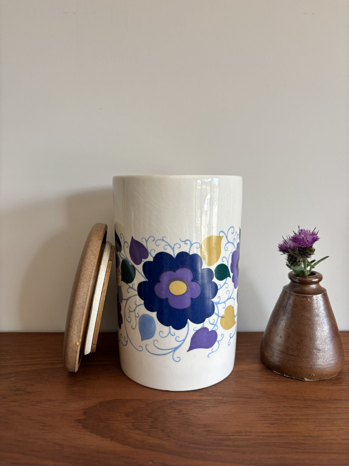 A white Crown Devon ceramic storage jar with a wooden lid, decorated with a blue, purple, and yellow floral pattern.