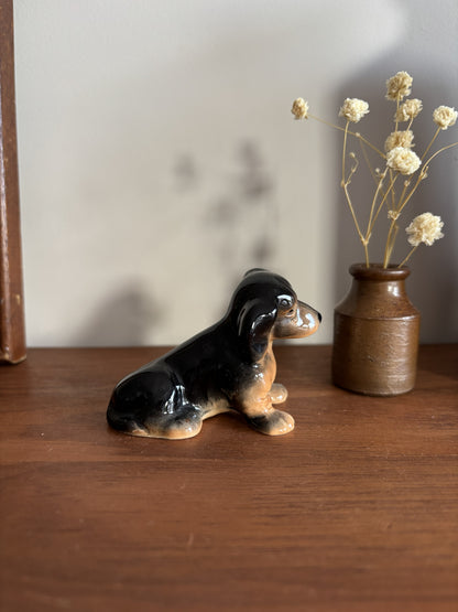 A glossy black and tan ceramic dachshund figurine sitting on a wooden shelf next to a small brown vase with dried cream flowers.