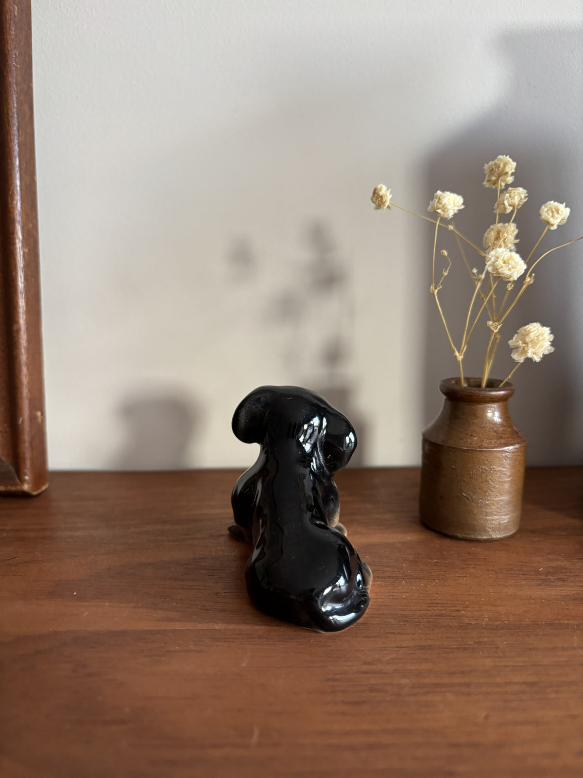A glossy black ceramic figurine of a dog sitting on a wooden table, next to a small brown ceramic vase holding dried cream-colored flowers against a neutral wall.